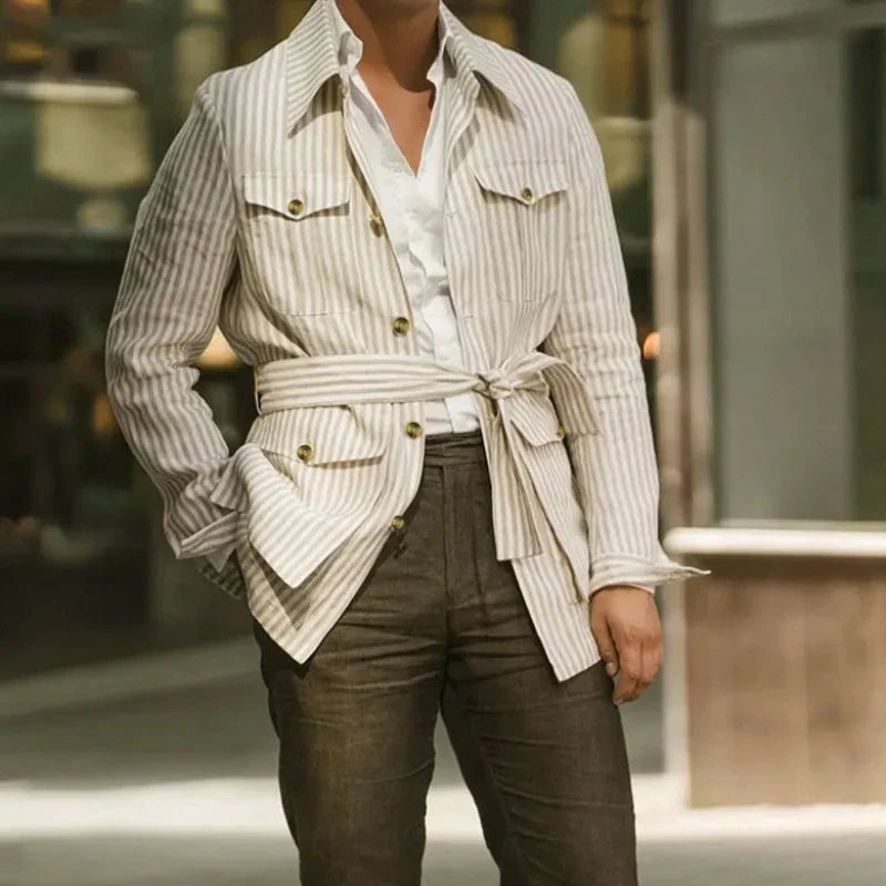 A person wearing a light-colored, striped jacket with an adjustable belt tied at the waist, white shirt, and brown trousers stands outdoors with one hand in their pocket. Their face is not visible.