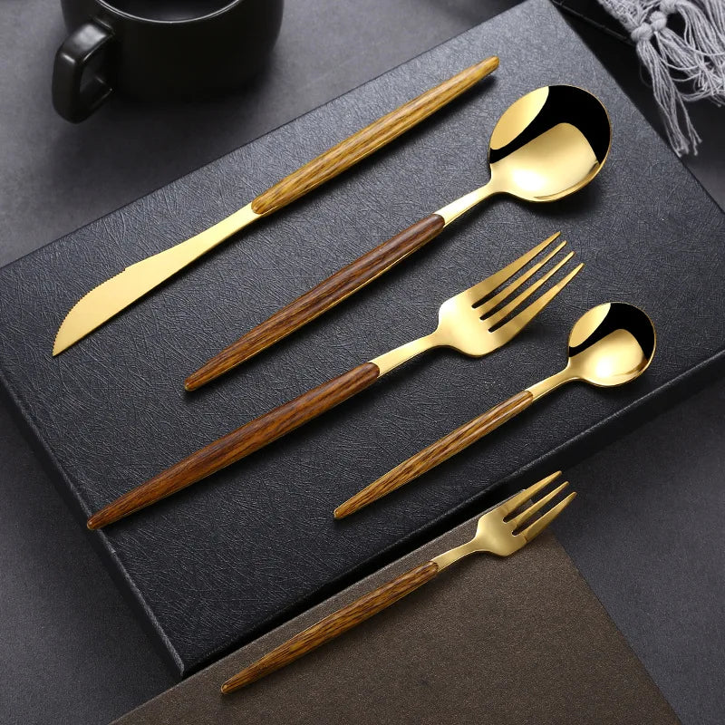 A gold plated flatware set with brown wooden handles, including a knife, tablespoon, fork, teaspoon, and dessert fork, is neatly arranged on a black textured surface with a cup and napkin in the background.