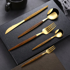 A gold plated flatware set with brown wooden handles, including a knife, tablespoon, fork, teaspoon, and dessert fork, is neatly arranged on a black textured surface with a cup and napkin in the background.