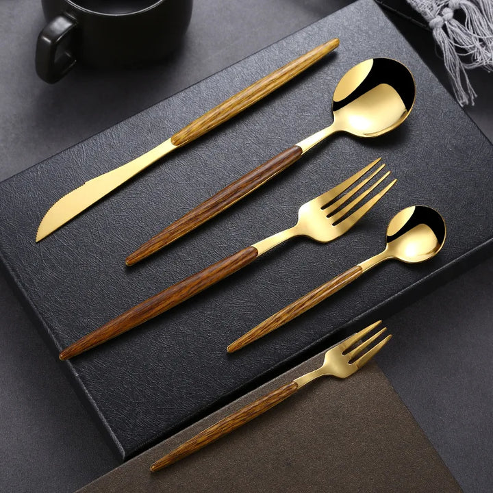 A gold plated flatware set with brown wooden handles, including a knife, tablespoon, fork, teaspoon, and dessert fork, is neatly arranged on a black textured surface with a cup and napkin in the background.