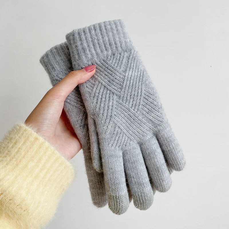 A hand wearing a pale yellow sweater holds a pair of luxurious, gray knit gloves with a ribbed cuff and criss-cross pattern, crafted from a cashmere blend, against a plain white background.