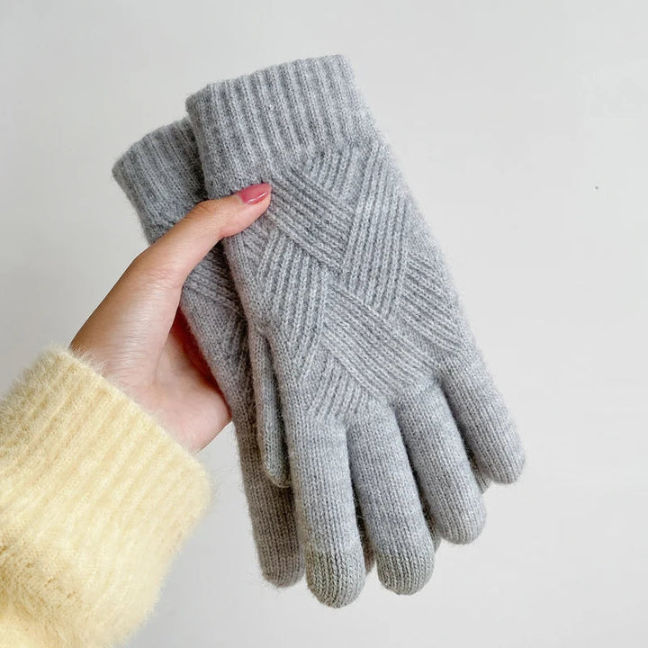 A hand wearing a pale yellow sweater holds a pair of luxurious, gray knit gloves with a ribbed cuff and criss-cross pattern, crafted from a cashmere blend, against a plain white background.
