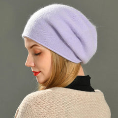 A woman in profile wears a lightweight, light purple knit beret and a beige sweater, with straight blond hair and red lipstick, against a plain gray background.