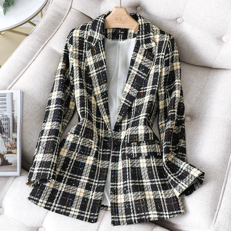 A black, white, and beige plaid Formal Blazer with front pockets is displayed on a beige upholstered chair. The stylish jacket coat hangs on a wooden hanger and features a light-colored inner lining.