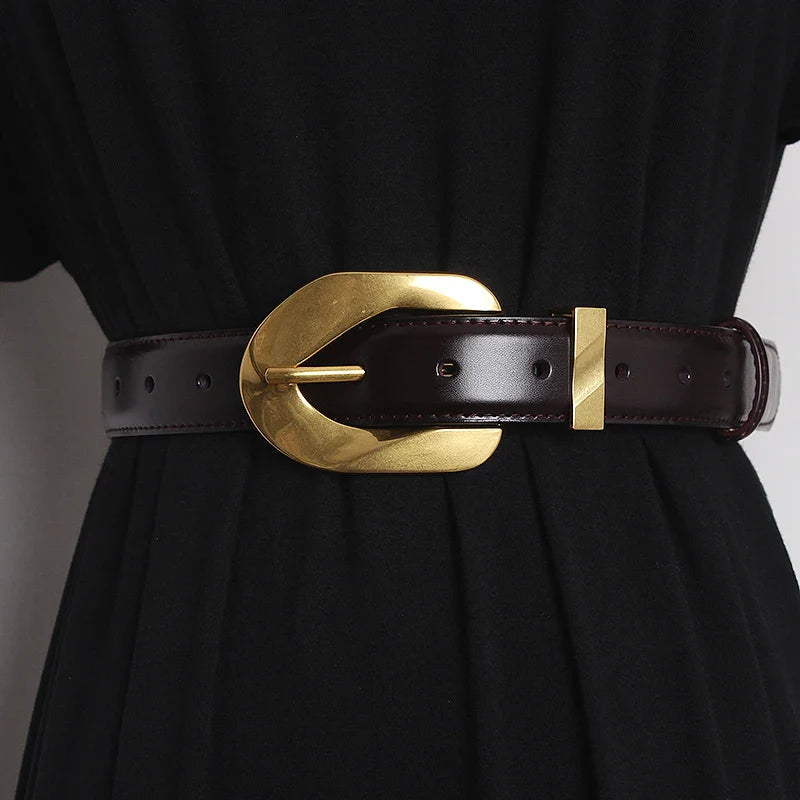 A person wearing a black dress with a wide, genuine leather belt featuring an oversized, oval-shaped gold buckle.