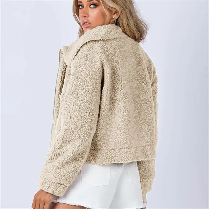 A woman with long, light brown hair wears a beige, cropped sherpa fur jacket and white denim shorts, standing sideways and looking over her shoulder against a plain light background—perfect for all season style.