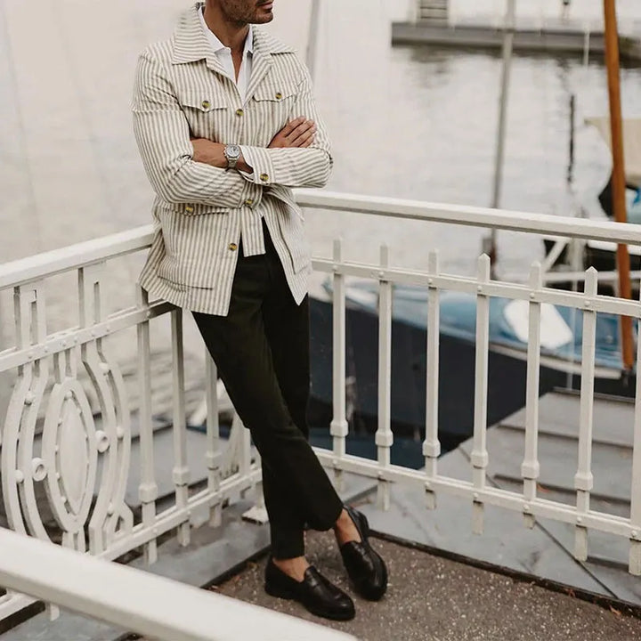 A man stands on a dock, leaning against a white metal railing. He wears a light pinstripe jacket in a tailored fit, dark pants, and black loafers. Boats and water are visible in the blurred background.