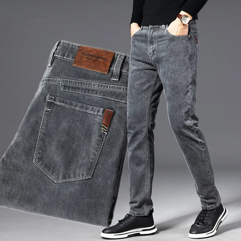 A person wearing slim-fit grey heavyweight denim jeans with hands in pockets, black sweater, watch, and black sneakers; a close-up of the jeans, showing the back pocket and leather label, is displayed beside them.