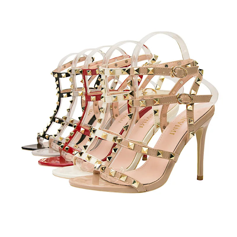 Three pairs of high-heeled sandals with studded embellishments are displayed in a row. The beige, red, and black shoes each feature gold-tone pyramid studs and adjustable buckle straps at the ankle.