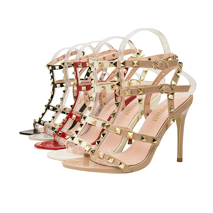 Three pairs of high-heeled sandals with studded embellishments are displayed in a row. The beige, red, and black shoes each feature gold-tone pyramid studs and adjustable buckle straps at the ankle.