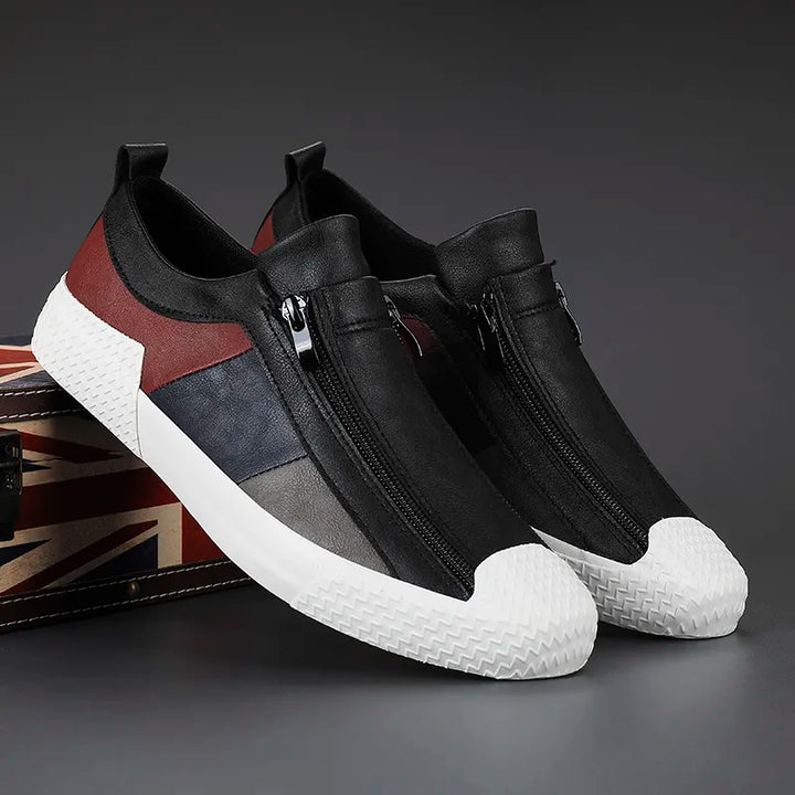 A pair of stylish slip-on sneakers with black, maroon, and gray color blocks, white textured rubber soles and toe caps, and a convenient zipper closure, displayed on a box with a British flag design.