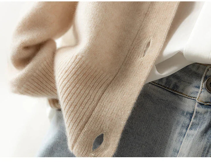 Close-up of a person wearing an ultra soft cashmere cardigan in beige, layered over a white shirt and paired with light blue high-waisted jeans; the 100% wool cashmere sweater features buttonholes along its edge.