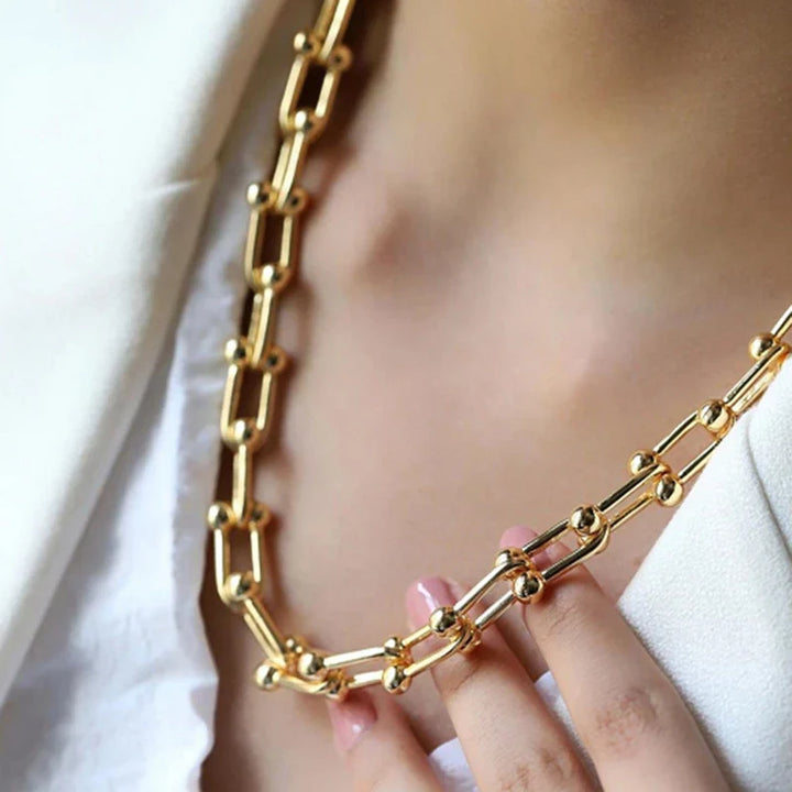A close-up of a person wearing a 316L stainless steel gold chain necklace with oval links and bead accents, dressed in a white top and blazer. The statement chain features an adjustable clasp, gently held between their fingers.