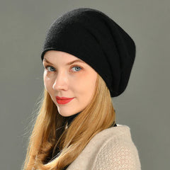 A woman with long blonde hair wears a loose, slouchy beanie and a beige textured sweater, looking over her shoulder and smiling slightly against a plain gray background.