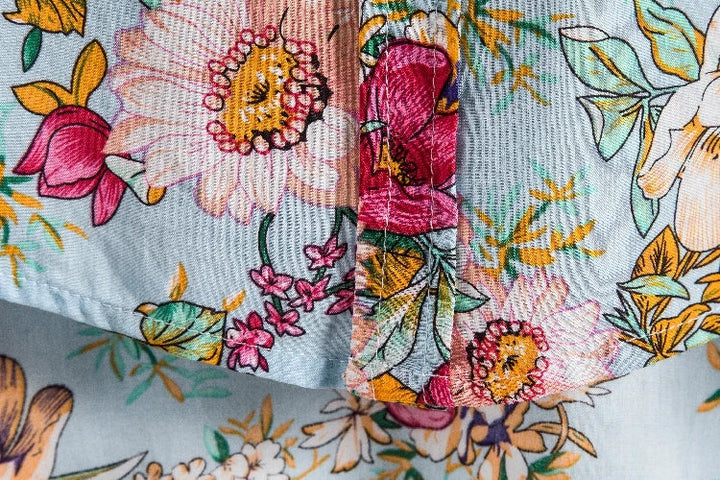 Close-up of colorful floral fabric, perfect for a vintage-inspired shirt, featuring large pink, yellow, and red flowers with green leaves on a light blue background. The textured cloth is slightly folded, highlighting the detailed pattern and stitching.