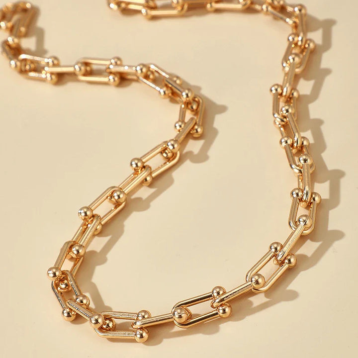 A statement chain necklace featuring elongated links and small gold beads at each joint, crafted from 316L stainless steel and displayed on a neutral beige background.