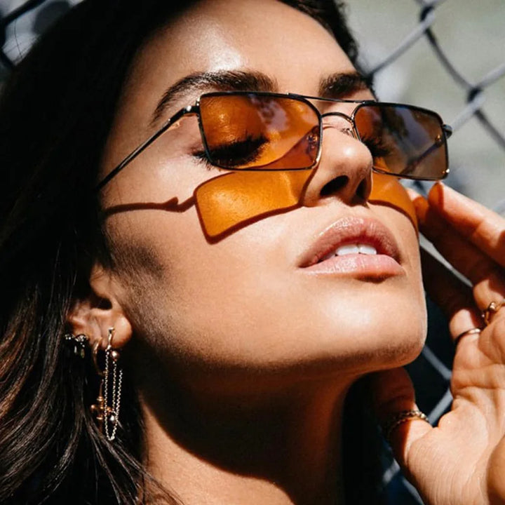 A woman with long brown hair wears rectangular, orange-tinted sunglasses crafted from lightweight alloy and gold hoop earrings. Tilting her head back with closed eyes, she touches her face as sunlight casts shadows on her UV400 protection lenses.