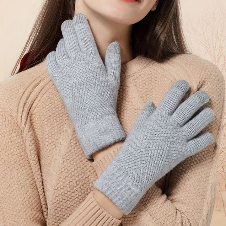 A woman wearing a beige knit sweater displays both hands, showing off luxurious, light gray knit gloves with a ribbed cuff and geometric pattern. Her hair is brown and loose, and she is smiling softly.