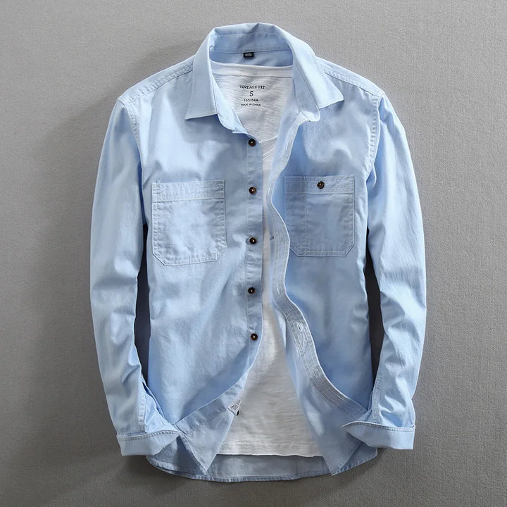 A light blue 100% cotton shirt with two chest pockets is worn open over a plain white T-shirt. The breathable shirt is displayed against a neutral gray background.