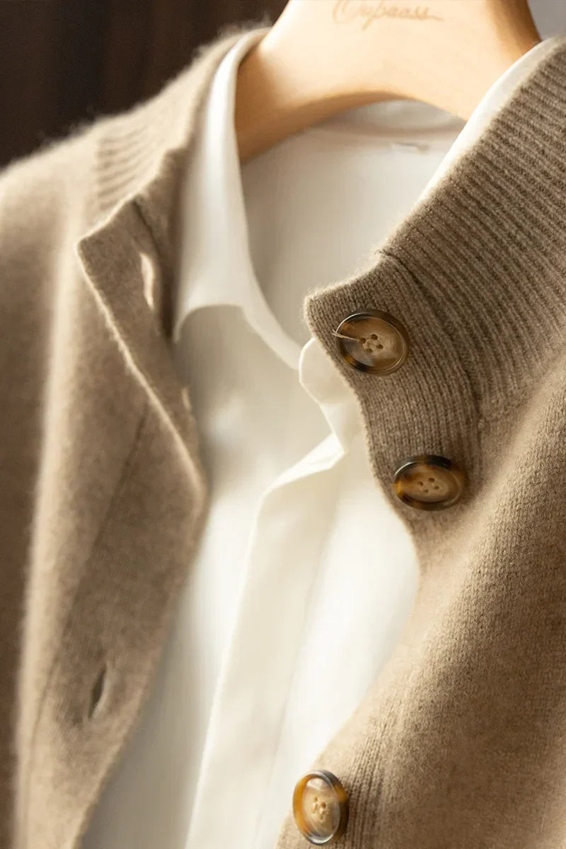 Close-up of an ultra soft cashmere cardigan in beige with large brown buttons, layered over a white collared shirt, both hung on a wooden hanger. Made from 100% wool cashmere for luxurious comfort.