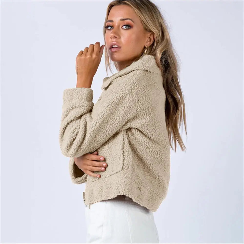 A woman with long blonde hair wearing a tan, textured sherpa fur jacket and white pants stands sideways, looking toward the camera against a plain light background.