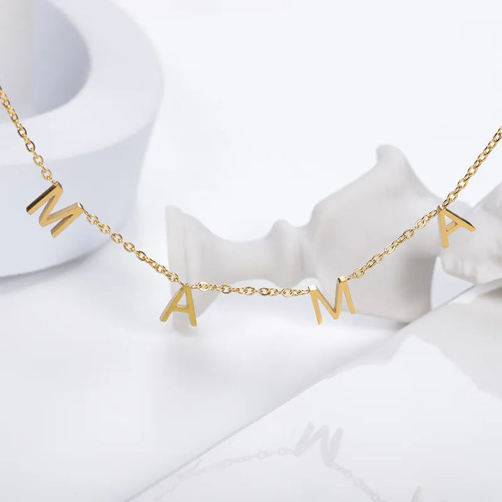 A gold minimalist jewelry piece, this stainless steel necklace features four dangling letter charms spelling MAMA, displayed against a white background with abstract white shapes.