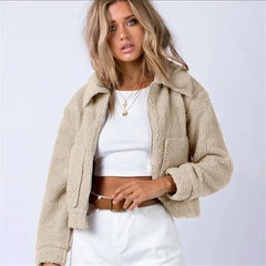 A woman with wavy, light brown hair wears a beige sherpa fur cropped jacket over a white crop top and high-waisted white pants. She accessorizes with layered necklaces and stands against a plain light background, showcasing an all-season look.