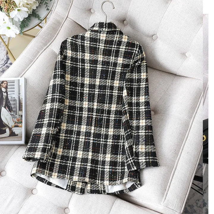 A black and white plaid formal blazer, perfect as an office work suit or stylish jacket coat, is displayed on a light-colored cushioned sofa with a hanger at the collar. A magazine and part of a bouquet are visible nearby.