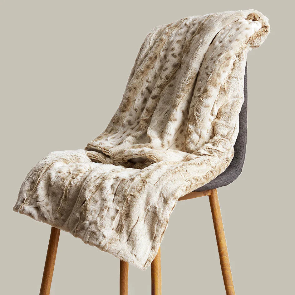 A beige ultra-soft luxurious blanket with a subtle pattern is draped over a modern gray chair with wooden legs, set against a plain light background.