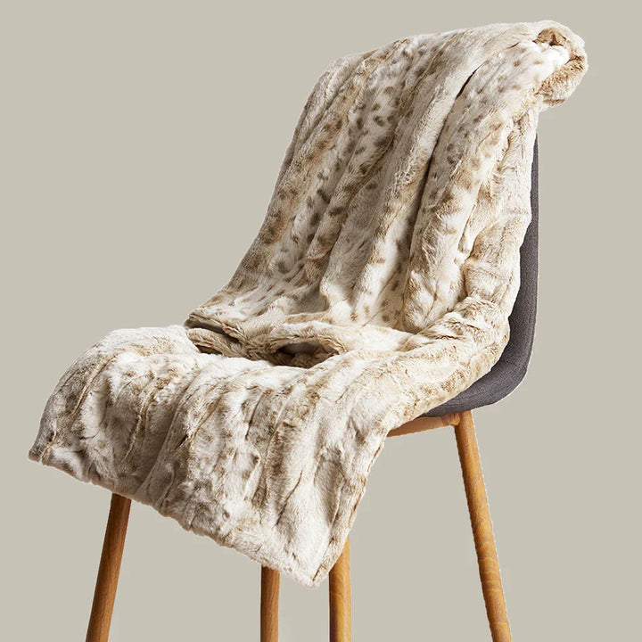 A beige ultra-soft luxurious blanket with a subtle pattern is draped over a modern gray chair with wooden legs, set against a plain light background.