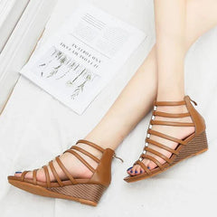 A person wearing brown strappy women shoes with wedge heels sits with legs crossed on a white surface next to an open magazine.