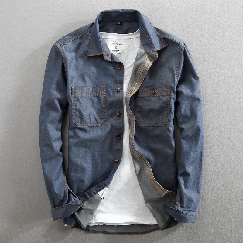 A blue denim 100% cotton shirt with two chest pockets is layered over a white t-shirt and laid flat on a light gray surface. The shirt is unbuttoned, and the sleeves are casually arranged.