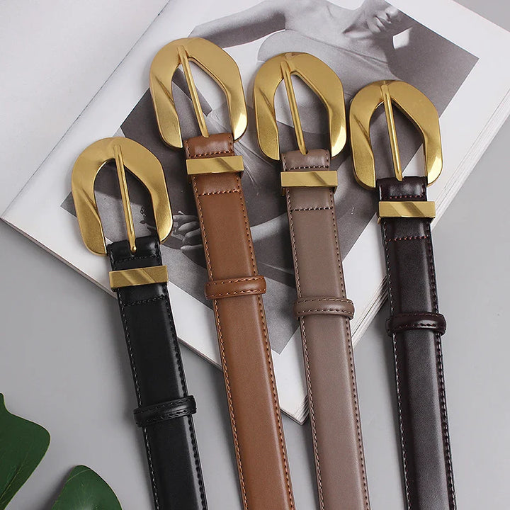 Four genuine leather belts in black, tan, taupe, and dark brown with oversized gold buckles are laid out on an open magazine featuring a grayscale fashion photo. Each adjustable belt adds bold style to any outfit.