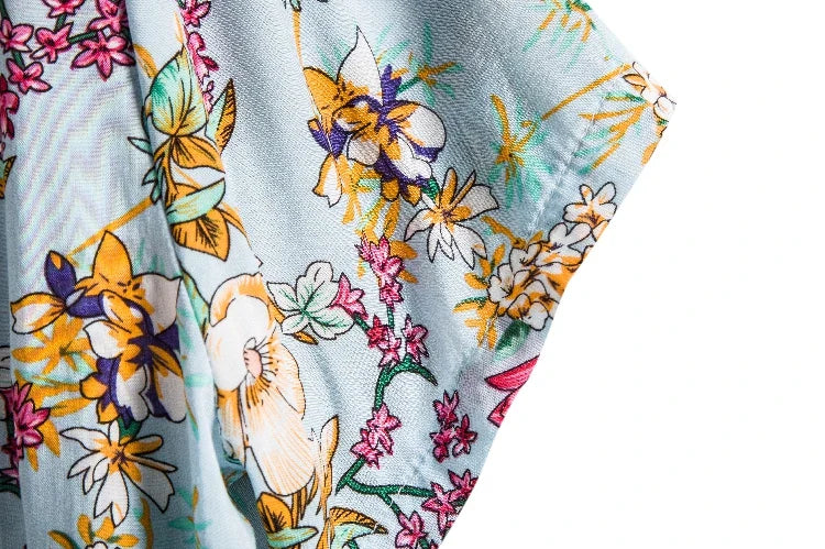 Close-up of a lightweight linen shirt in light blue, adorned with a colorful floral pattern featuring yellow, white, and purple flowers with green stems and small pink blossoms, set against a white background.