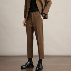 A person wearing mens slim casual trousers in premium fabric, paired with black socks and dress shoes, stands on a wooden floor with hands in pockets. Only the lower half is visible against a plain beige background.