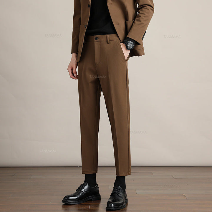 A person wearing mens slim casual trousers in premium fabric, paired with black socks and dress shoes, stands on a wooden floor with hands in pockets. Only the lower half is visible against a plain beige background.