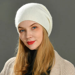 A woman with long blonde hair wears a slouchy beanie, a beige sweater, and a black turtleneck. She is looking at the camera and smiling slightly against a plain gray background.