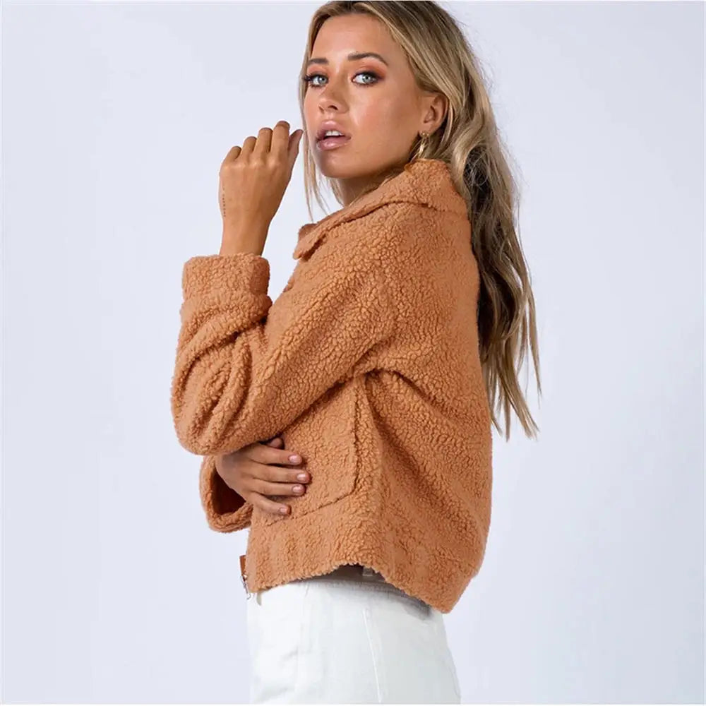 A woman with long blonde hair wears a tan, cropped sherpa fur jacket and white pants, posing sideways against a plain light background.