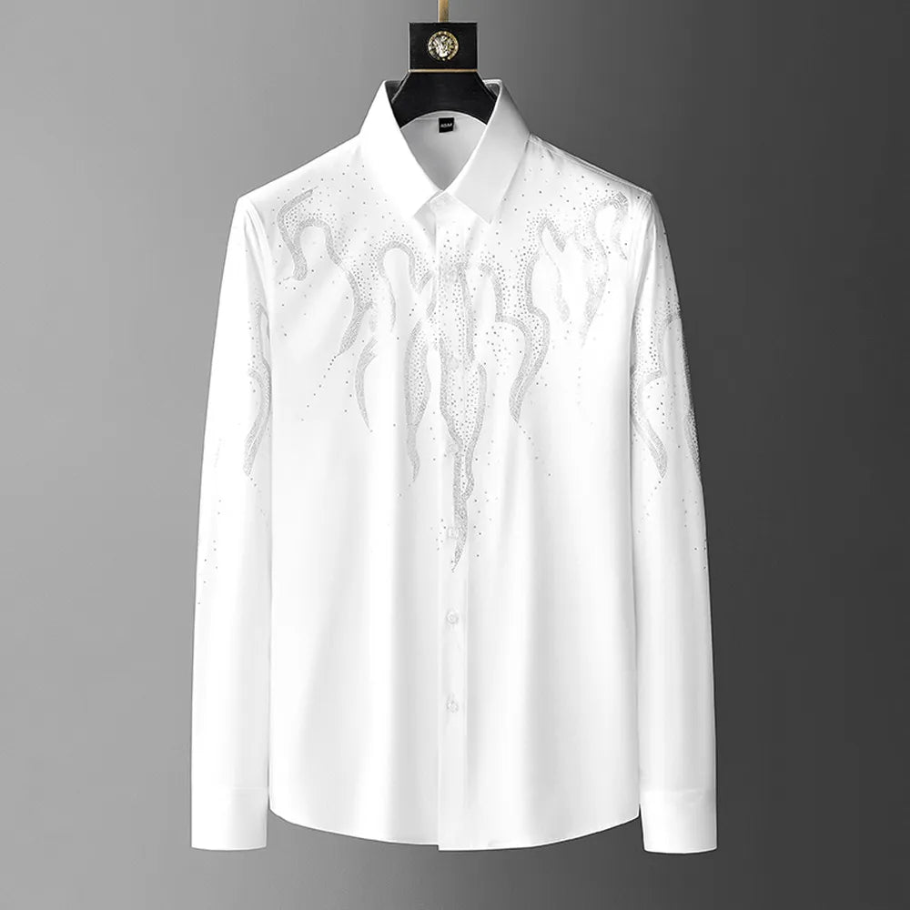 A white tailored fit button-up shirt with a pointed collar, featuring sparkling rhinestones arranged in a flame-like design on the chest and shoulders, hangs stylishly on a black hanger.