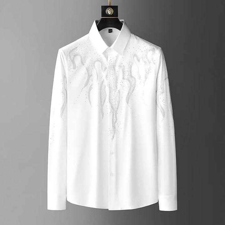 A white tailored fit button-up shirt with a pointed collar, featuring sparkling rhinestones arranged in a flame-like design on the chest and shoulders, hangs stylishly on a black hanger.