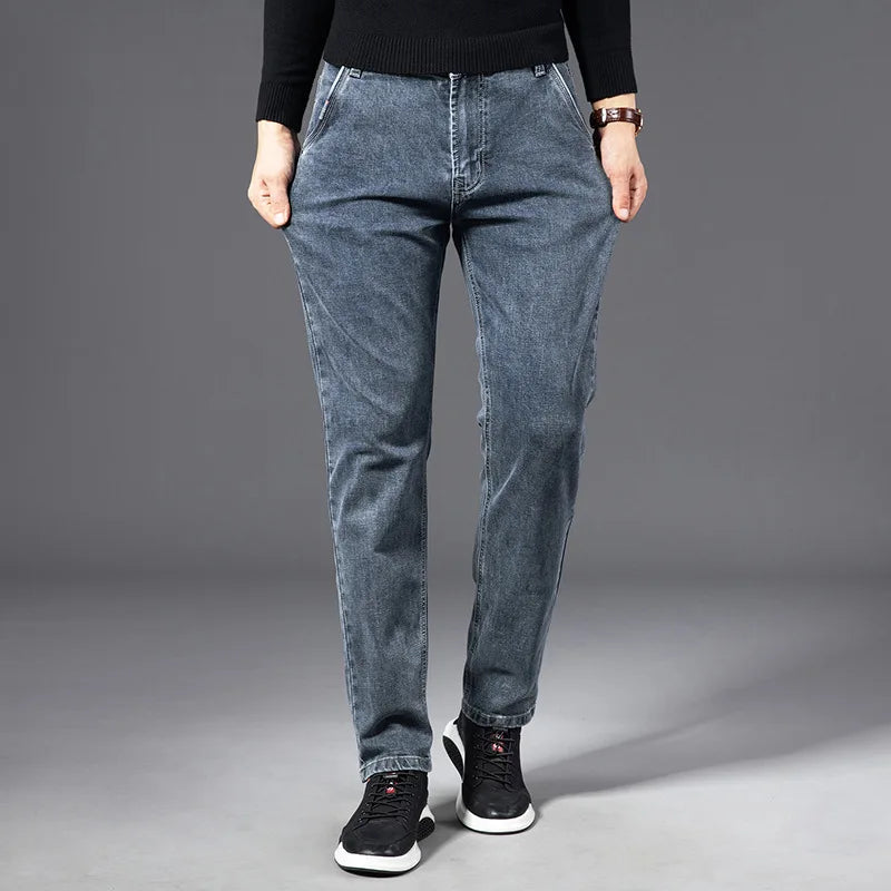 A person wearing a black sweater, straight-leg blue jeans, and black sneakers stands with hands in pockets against a plain gray background.