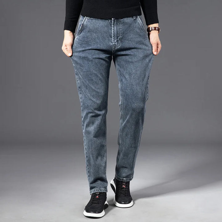 A person wearing a black sweater, straight-leg blue jeans, and black sneakers stands with hands in pockets against a plain gray background.