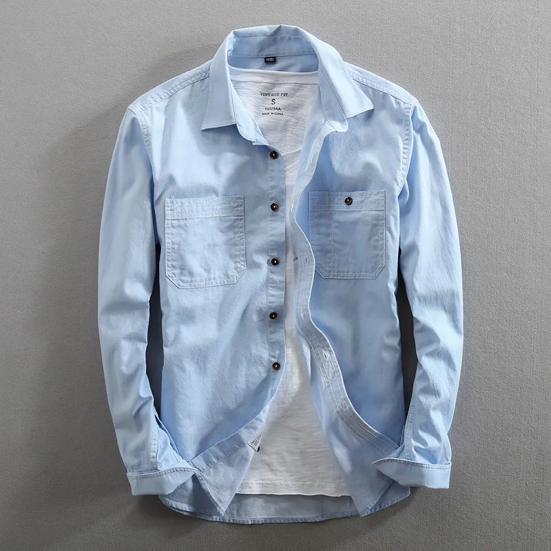 A light blue 100% cotton shirt with two chest pockets, displayed open over a white t-shirt, laid flat against a gray background.
