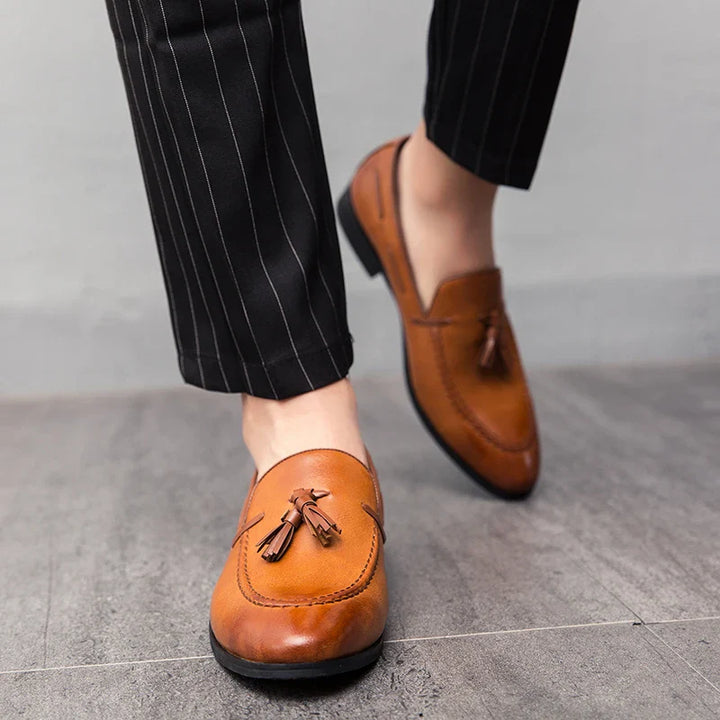 Close-up of a person wearing genuine leather mens loafers with tassels and black pinstripe pants, standing on a gray tiled floor.