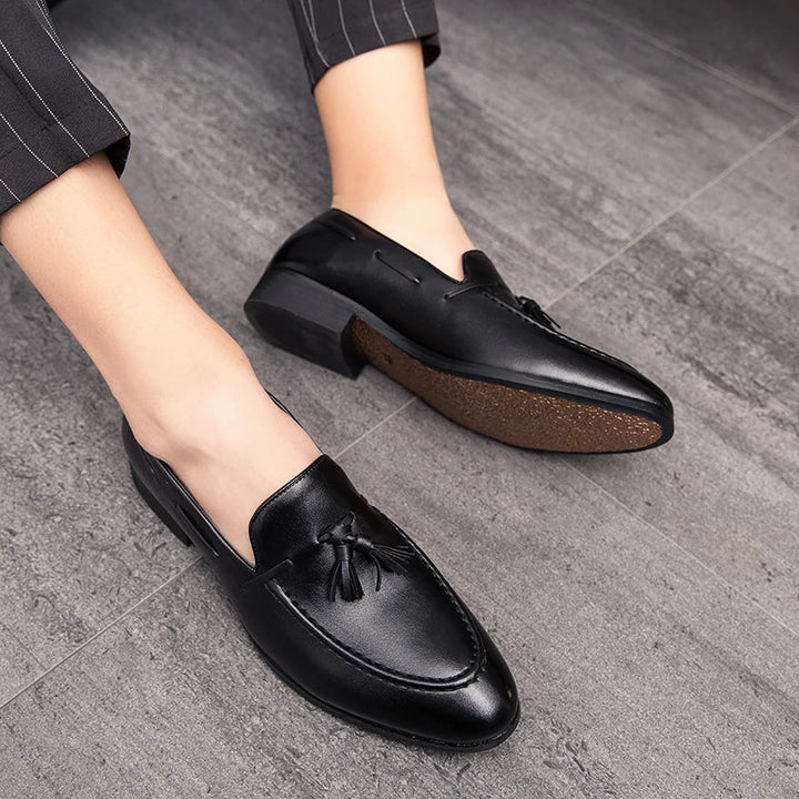 A person wearing black genuine leather tassel men’s loafers with pinstriped pants, standing on a gray tiled floor.