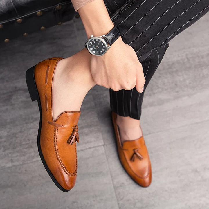 A person wearing mens loafers made of genuine leather with brown tassels and black pinstripe pants sits on a chair, one leg crossed over the other, showing a wristwatch on their left wrist.