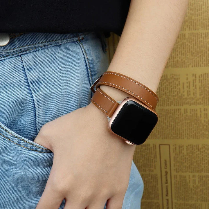 A person wearing blue jeans and a black top sports an Apple Watch with a genuine leather double wrap design band, its adjustable sizing offering comfort as their hand rests casually by their side.