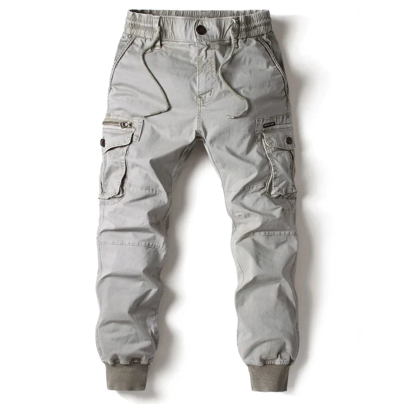Light gray cargo jogger pants featuring street style with an elastic waistband, drawstring, side cargo pockets with button flaps, front zipper pocket, and ribbed cuffs at the ankles, laid flat on a white background.