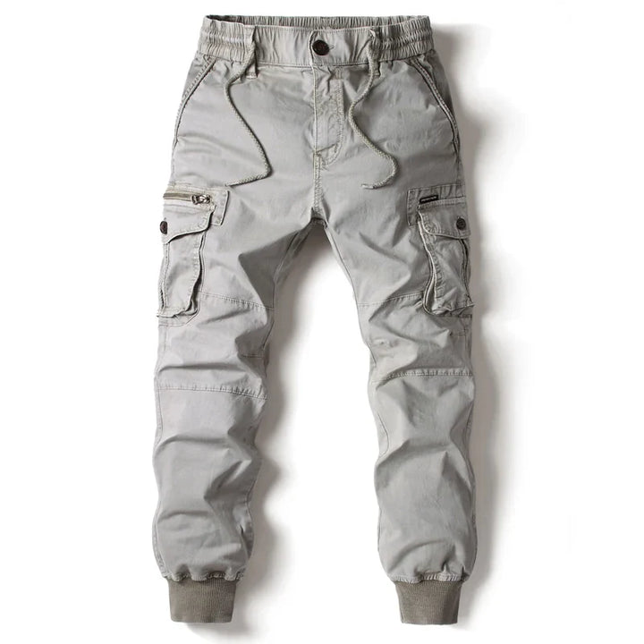 Light gray cargo jogger pants featuring street style with an elastic waistband, drawstring, side cargo pockets with button flaps, front zipper pocket, and ribbed cuffs at the ankles, laid flat on a white background.