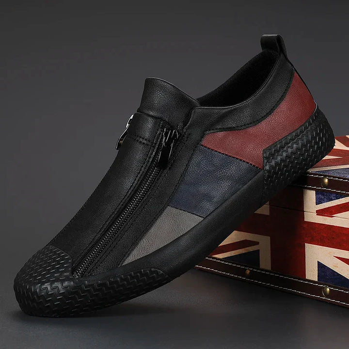 A stylish black slip-on sneaker with a zipper closure, featuring dark red, navy, and gray panels. The cushioned sole adds comfort as the shoe is propped against a suitcase decorated with the British flag.